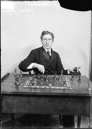 Chess player Frank Marshall, holding a chess piece and a cigar, sitting behind a table in front of a light-colored backdrop in a room - Chicago Daily News, Inc.