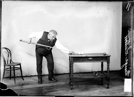 Billiards player George Sutton standing to left of a desk, aiming the cue at a cue ball, wooden chair is visible to the left of Sutton - Chicago Daily News, Inc.