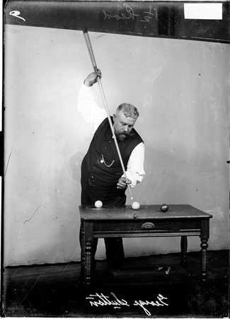 Billiards player George Sutton standing behind a desk, holding a cue almost vertically and aiming it at a cue ball set up on the desk - Chicago Daily News, Inc.