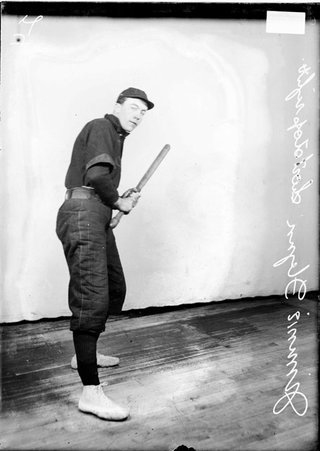 Indoor baseball right shortstop, Jimmie Flynn, holding a bat and standing with his right side to the camera in front of a backdrop - Chicago Daily News, Inc.