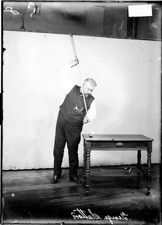 Billiards player George Sutton standing at corner of a desk, holding a cue almost vertically and aiming at a cue ball on the desk - Chicago Daily News, Inc.