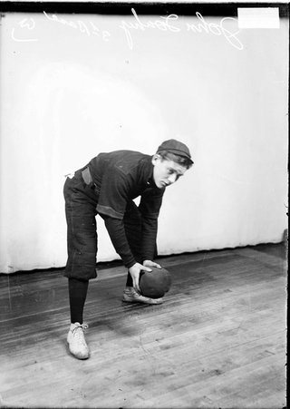 Indoor baseball third baseman, John Touhy, bending forward and holding a large, soft ball in his hands, in front of backdrop in a room - Chicago Daily News, Inc.