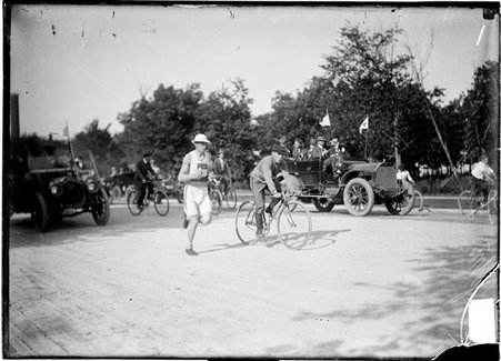 Athlete John Lindquist, running on a road during Illinois Athletic Club Marathon, auto with flags on it is visible in the background - Chicago Daily News, Inc.