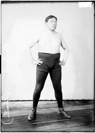 Native American wrestler War Eagle, standing facing the camera with his arms resting on his hips in front of a backdrop, wearing trunks - Chicago Daily News, Inc.