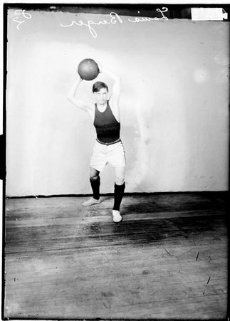 Hull House basketball player Louis Berger, holding a basketball above his head with both hands standing in front of a backdrop - Chicago Daily News, Inc.