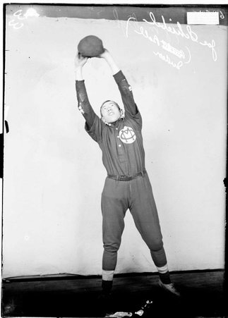 Indoor baseball player Joe Schiebel, holding a ball with both hands stretched above his head standing in front of a backdrop in a room - Chicago Daily News, Inc.
