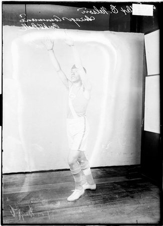 Chicago Commons basketball player Alex E. Nelson, reaching both hands above his head standing in front of a backdrop in a room - Chicago Daily News, Inc.