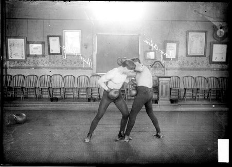 Boxers Tony Caponi and Hugo Kelly, standing on a large rug, sparring, Kelly is jabbing left into Caponi's stomach - Chicago Daily News, Inc.
