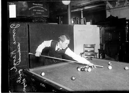 Billiards player Jerome Keogh, aiming a cue at the cue ball leaning across the pool table - Chicago Daily News, Inc.