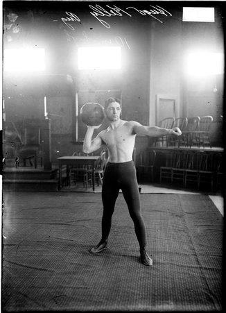 Boxer Hugo Kelly, standing on a rug in a room and holding a medicine ball in his right hand over his shoulder - Chicago Daily News, Inc.