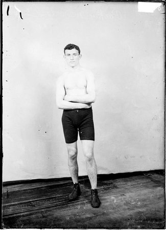Pugilist, Packey McFarland, looking at the camera with his arms crossed at his waist in front of a light colored background in a room - Chicago Daily News, Inc.