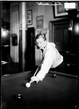 Billiards player Thomas Hueston, aiming a cue at the cue ball leaning across the billiards table in a room - Chicago Daily News, Inc.