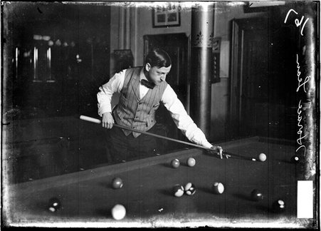 Billiards player Horace Lean, aiming a cue at the cue ball, standing behind a pool table in a room - Chicago Daily News, Inc.