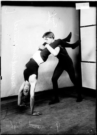 Wrestlers, Jahn holding Armbruster upside down by his legs, Armbruster supporting himself with his arms in front of a light colored background in a room - Chicago Daily News, Inc.