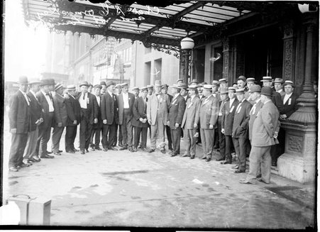 White Sox owner Charles Comiskey, standing with a group of men wearing ribbons on their jackets in front of a building, light exposure - Chicago Daily News, Inc.