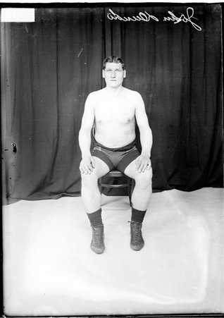 Wrestler John Dundo, wearing wrestling tights, sitting in front of a dark backdrop in a room - Chicago Daily News, Inc.