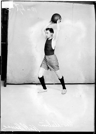 Hull House basketball player and captain George Dubin, holding a basketball above his head, standing in front of a backdrop - Chicago Daily News, Inc.