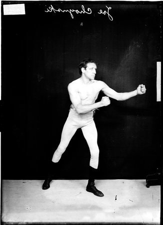 Pugilist Joe Choynski, wearing boxing tights and facing to right of camera, standing in a boxing stance in front of a backdrop - Chicago Daily News, Inc.