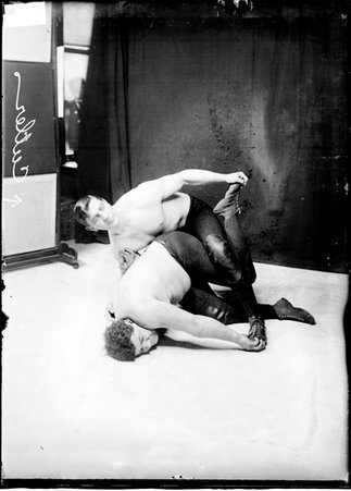 Wrestler C. Cutler kneeling behind an unidentified wrestler and holding him by the arm and toe in front of a dark backdrop - Chicago Daily News, Inc.