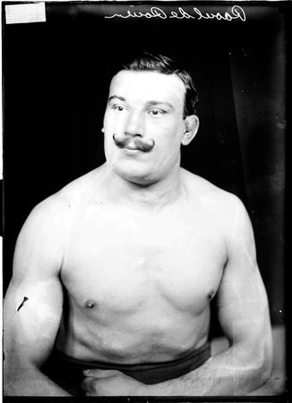 French wrestler Raoul De Rouen, holding his forearms across his abdomen, flexing his muscles, sitting shirtless in front of a backdrop - Chicago Daily News, Inc.