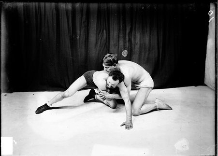 Unidentified wrestler holding another unidentified wrestler with his hands clasped around his arm, kneeling in front of a dark backdrop - Chicago Daily News, Inc.