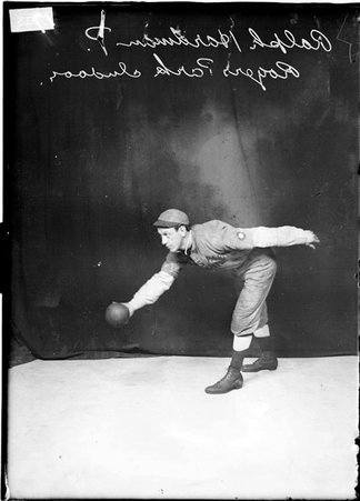 Rogers Park indoor baseball player Ralph Hardman, leaning forward, holding a baseball in front of him, standing in front of a backdrop - Chicago Daily News, Inc.