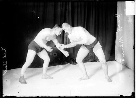 Wrestlers, Yussiff Mahout and Fred Beell facing one another, standing in ready positions in front of a dark backdrop - Chicago Daily News, Inc.