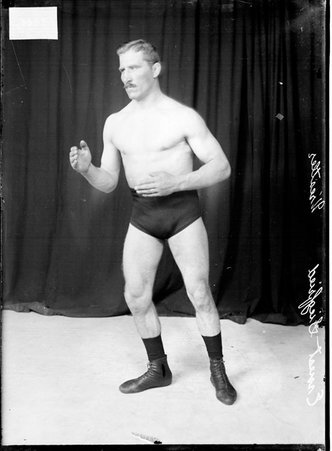 Wrestler Ernest Siegfried, holding his hands in front of him, standing in an athletic position in front of a dark backdrop - Chicago Daily News, Inc.