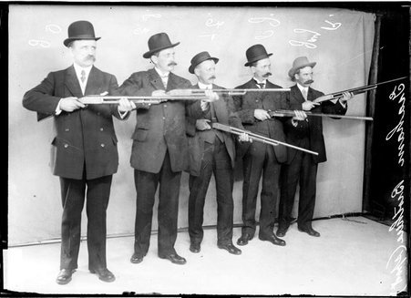 Trapshooters the Graham brothers, aiming rifles to the right of the camera, standing in front of a light-colored backdrop - Chicago Daily News, Inc.