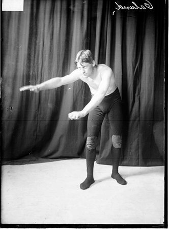 Wrestler Oslund, leaning forward and extending his arms in front of him, standing in front of a dark backdrop - Chicago Daily News, Inc.