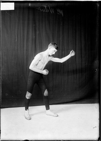 Wrestler William Waters wearing tights and holding his left arm out in front of him, standing in front of a dark backdrop - Chicago Daily News, Inc.