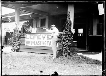 Shooter E. B. Shegren aiming a rifle, standing in front of a low building at the Chicago Gun Club - Chicago Daily News, Inc.