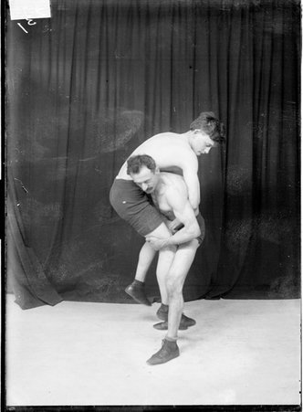 Unidentified wrestler carrying another unidentified wrestler over his right shoulder, standing in front of a dark backdrop - Chicago Daily News, Inc.