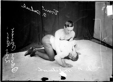 Wrestlers, W. R. Milchewski and Emil C. Schalk, Davis Square wrestlers, Schalk is kneeling over Milchewski holding his head to floor in front of dark backdrop - Chicago Daily News, Inc.