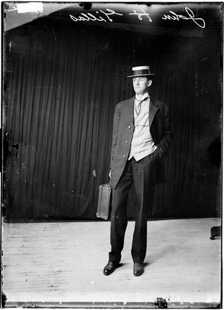 Athlete, John H. Gillas standing against dark backdrop wearing suit and hat, carrying suitcase - Chicago Daily News, Inc.