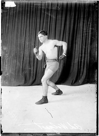 Pugilist, Ad. Wolgast, standing with knees bent, right fist forward, wearing boxing gloves against dark backdrop - Chicago Daily News, Inc.