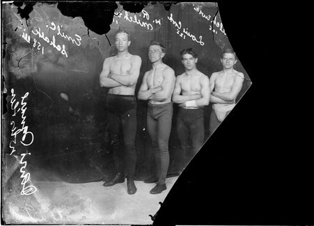 Wrestlers, Louis Cook, Elmer Milchewski, W. R. Milchewski, and Emil C. Schalk, Davis Square wrestlers, standing with arms crossed in front of dark backdrop - Chicago Daily News, Inc.