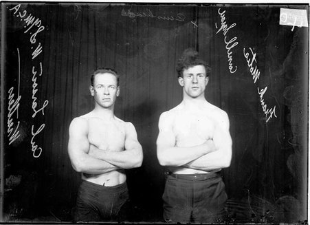Wrestlers, Carl Larson, West Side YMCA, and Frank Motis, Cornell Square, standing with arms crossed on their chests in front of dark background - Chicago Daily News, Inc.