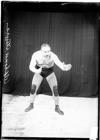 Wrestler Alfonse Steurs posing in a wrestling stance with his arms pointing to the right of the image in front of a dark colored background - Chicago Daily News, Inc.