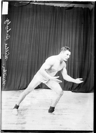 Wrestler Willie Schultz standing in a wrestling stance, lunging to the side on his left leg, in front of a dark colored background - Chicago Daily News, Inc.