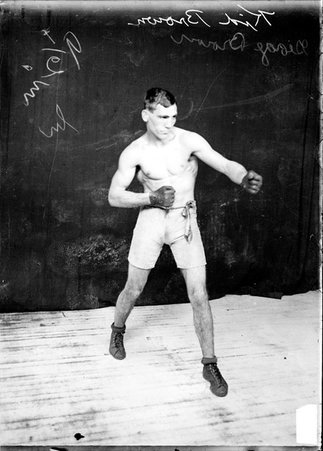 Boxer Kid Brown standing in a boxing stance, fists pointed to the right of the image, in front of a dark colored background - Chicago Daily News, Inc.