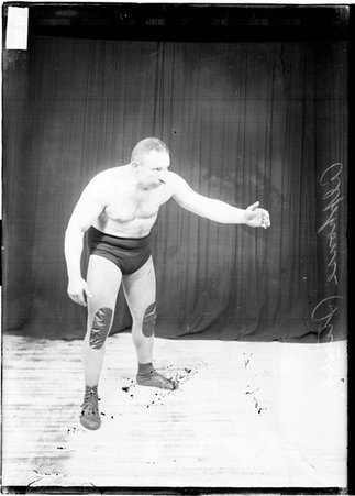Wrestler Alfonse Steurs in a wrestling stance with his left arm out in front of him standing in front of a dark colored background - Chicago Daily News, Inc.