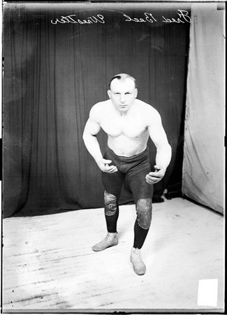 Wrestler Fred Beell standing in a typical wrestling stance, bent slightly forward, in the corner of light and dark colored backgrounds in a room - Chicago Daily News, Inc.