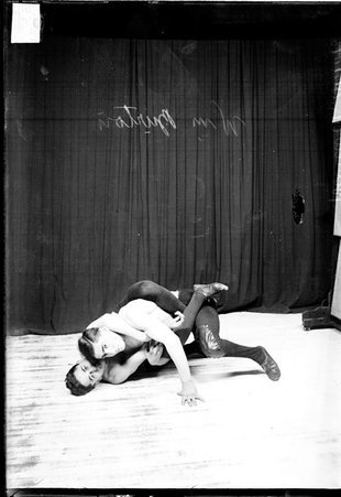 Wrestlers William Burton and Leo Kallis posing, performing a wrestling maneuver on the floor in front of a dark colored background - Chicago Daily News, Inc.