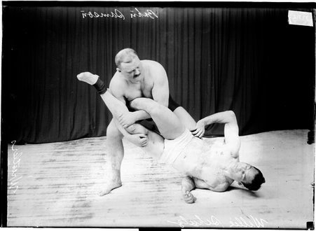 Wrestlers Samson holding Schultz's legs in a wrestling maneuver in front of a dark colored background - Chicago Daily News, Inc.