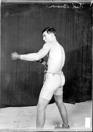 Boxer Kid Brown standing in a boxing stance with his back to the camera in front of a dark colored background He wearing leather gloves on his hands. - Chicago Daily News, Inc.
