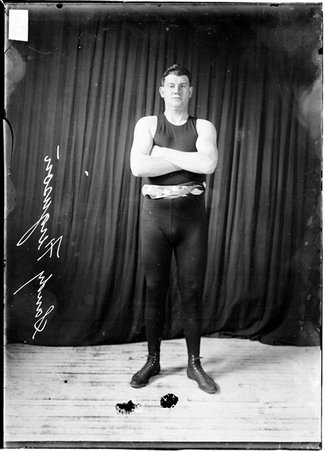 Fighter, Sandy Ferguson standing with arms crossed against dark backdrop, wearing Am. flag around waist - Chicago Daily News, Inc.