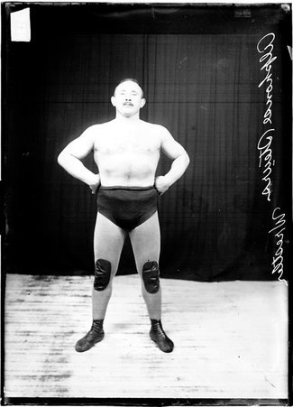 Wrestler Alfonse Steurs standing with his hands on his hips in front of a dark colored background - Chicago Daily News, Inc.
