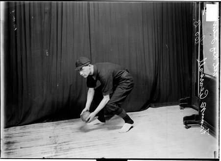 Seventh Regiment indoor baseball player Frank Pearsall stooping to pick up a ball standing in front of a dark colored background - Chicago Daily News, Inc.