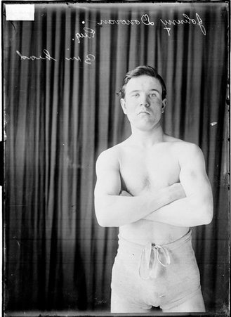 Boxer Johnny Donovan standing with his arms crossed on his chest in front of a dark colored background - Chicago Daily News, Inc.